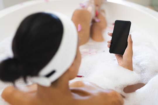 Mixed race woman in bathroom relaxing in bath and using smartphone