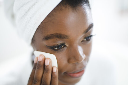 Smiling african american woman in bathroom cleansing her face with cotton pad for skin care