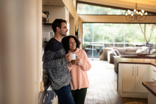Happy diverse couple in kitchen embracing drinking coffee and smiling