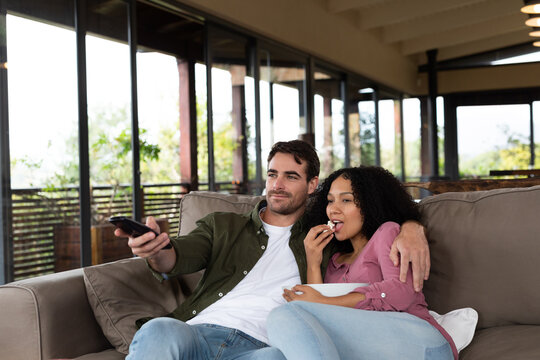 Happy diverse couple sitting on sofa in living room embracing and watching tv