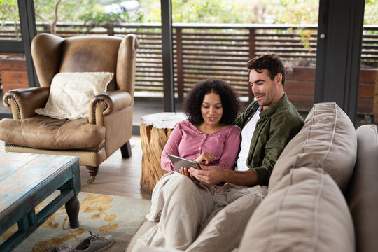 Happy diverse couple sitting on sofa in living room using tablet