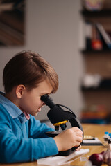 Distance online education, internet learning. First grade boy studying at home using microscope, making notes, biology online lesson