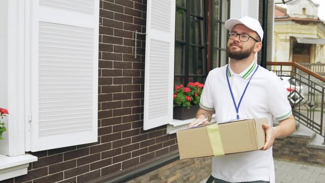 Motion of young cheerful bearded postman in white cap and casual outfit carrying box, ringing at doorbell. Tattooed deliveryboy with badge comparing house number with data in folder. Delivery concept.