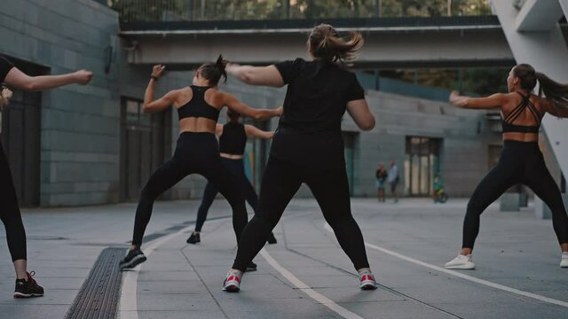 Back view group of girls under the guidance of a trainer perform a set of aerobics exercises and dance zumba. Outdoor morning warm-up