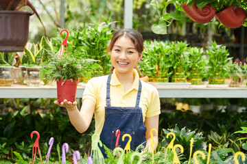 Portrait of cheerful young greenhouse worker showing fern plant in hanging pot