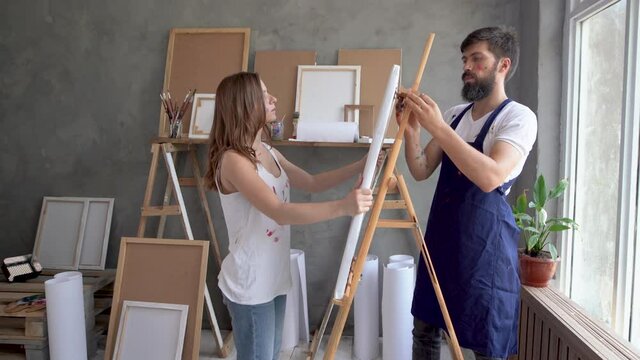 A man and a woman in an art studio walk up to the easel and fix the canvas.