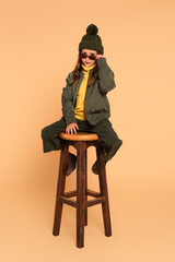 trendy kid looking at camera over sunglasses while sitting on high wooden stool on beige