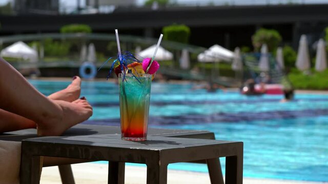 Woman relaxing at the side of a pool with a colorful cocktail in a tall glass alongside her on a table in a close up on her legs on a recliner chair