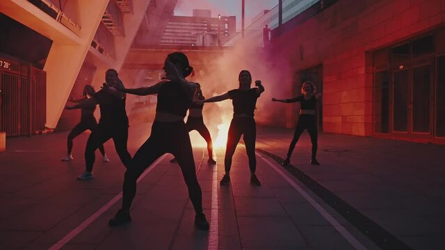 Silhouettes of six beautiful girls dancing zumba at night on a city street against the background of a smoke bomb or a torch. Performance and women's movement concept