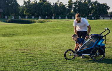 Fototapeta na wymiar Father playing and communicate with his child in baby stroller while standing on a green lawn on a sunny day. Togetherness father and son, father's day, parenthood