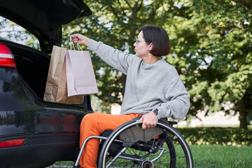 Woman sitting at her wheelchair and opening car trunk and putting shopping bags inside