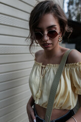 Cool portrait of fashionable woman with stylish round sunglasses in yellow blouse top walks near wall on street