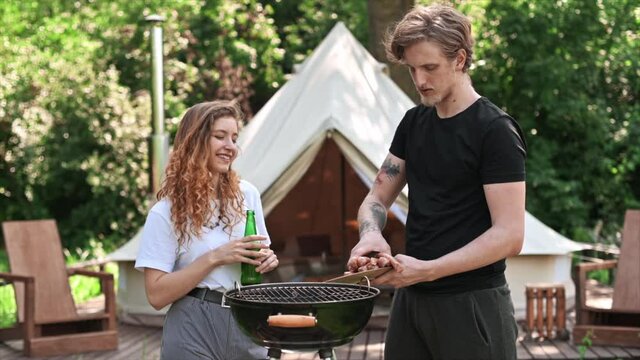 Young couple frying meat on the grill and drinking beer. Greenery around. Glamping. Slow motion