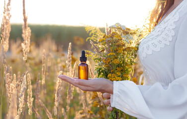 A woman in a white dress holding a bottle with a dropper in the background of herbs, a field...