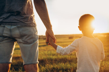Father's and his son holding hands at sunset field. Dad leading son over summer nature outdoor....