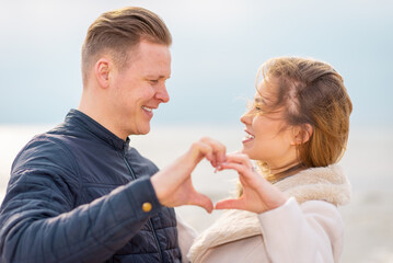 Two young couple is making heart, looking at each other and smiling while standing at the beach.