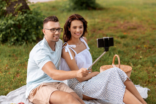 leisure and people concept - happy couple having picnic and taking picture with smartphone on selfie stick at summer park