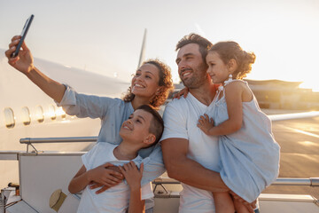 Happy tourists, beautiful family taking selfie while standing together outdoors ready for boarding the plane at sunset. Vacation, parenthood, traveling concept - obrazy, fototapety, plakaty