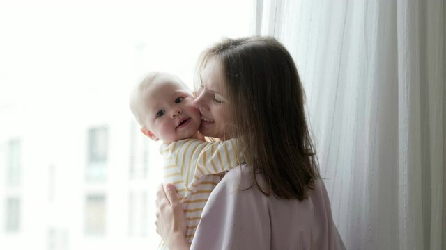Young beautiful mother embracing her little baby. Mom and daughter hugging together sitting on windowsill and looking out through window. family love