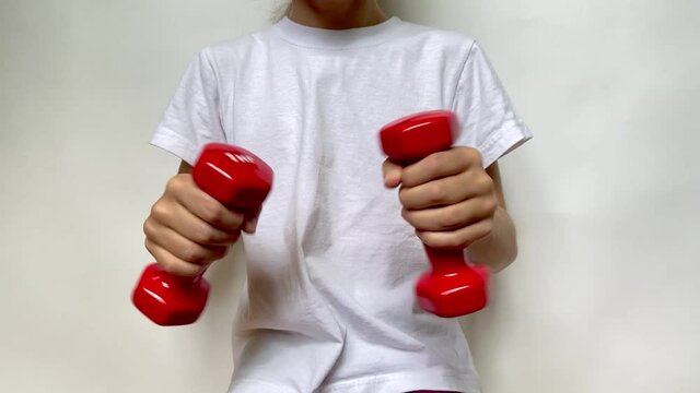 A girl in a white T-shirt slowly moves two red dumbbells weighing 1 kg 