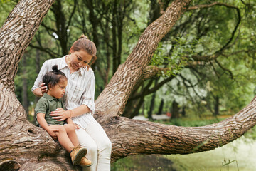 Portrait of happy young mother playing with son in park while sitting on tree branches, copy space