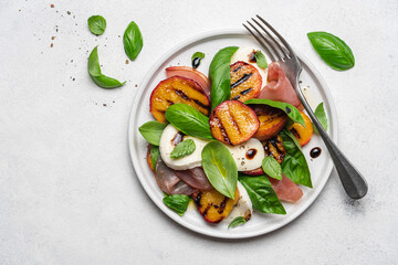 Grilled peach salad with mozzarella, prosciutto ham, basil, olive oil and balsamic. Fresh gourmet salad in plate on white stone background