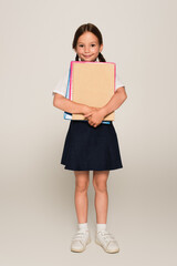 full length view of schoolgirl with notebooks looking at camera on grey