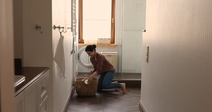Young Indian housewife do regular housework, carries laundry wicker basket walk along laundry room puts bath towels into washing machine busy in routine household in modern cozy home, cleaning concept