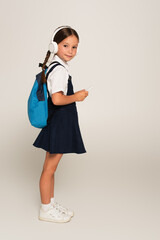 full length view of schoolgirl in headphones holding mobile phone on grey