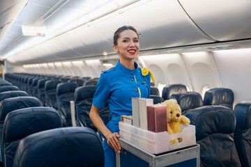 Cheerful female air hostess in bright blue uniform smiling at camera while leading trolley cart...