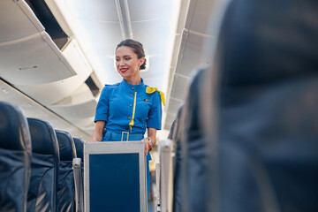 Smiling female cabin attendant leading trolley cart through empty plane aisle. Travel, service,...