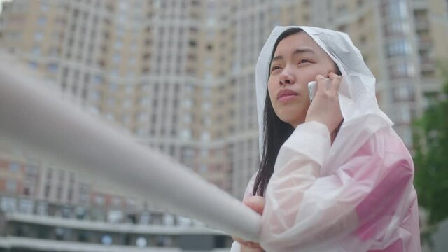 Upset asian woman in raincoat talking on phone in rainy city, calling taxi