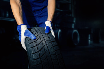 mechanic holding car tire on dark workshop background with copy space. tyre service - obrazy, fototapety, plakaty