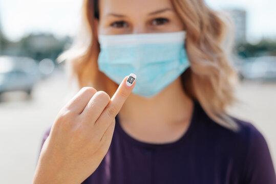 Close-up of young woman in medical face mask shows qr code manicure on finger on street outside, confirmation of coronavirus vaccination. New normal concept, pass by qr code