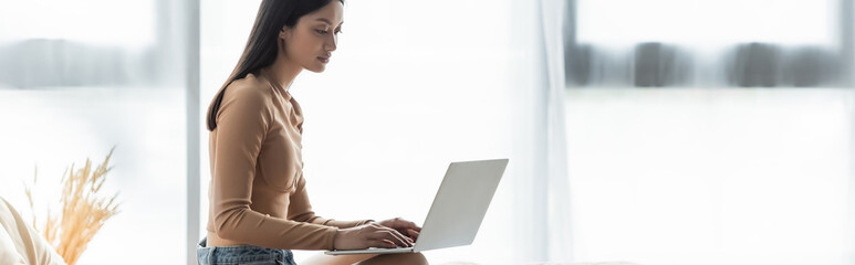 Naklejka premium pretty asian freelancer typing on laptop near window at home, banner.