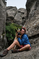 Female tourist on mountain hike in blue jacket and boots. Pretty young Caucasian woman is sitting on rock and hugging dog. Hiking in mountains with German Shepherd.