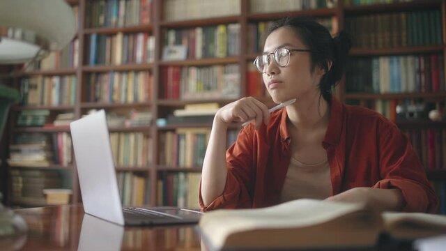 Asian student thinking over essay plot, working with books and pc at the library