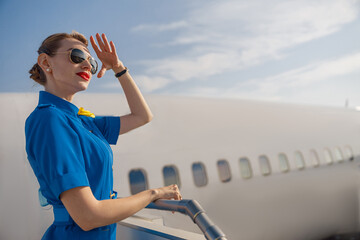 Portrait of elegant air stewardess in blue uniform and sunglasses holding hand near her head and...