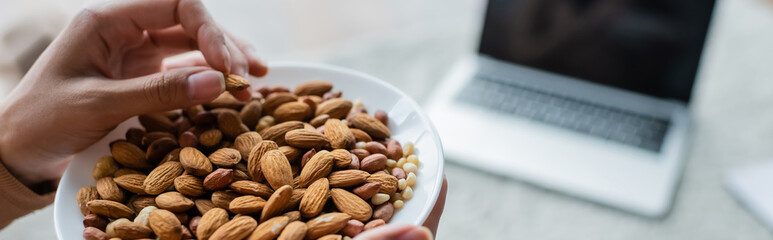 partial view of woman with bowl of almonds near blurred laptop with blank screen, banner.