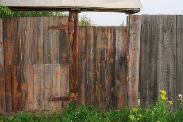 A gate with metal elements in a board fence near an old village house in the Urals (Russia) in summer. close-up. The texture of the old fence with spots. Green grass and cloudy sky serve as background