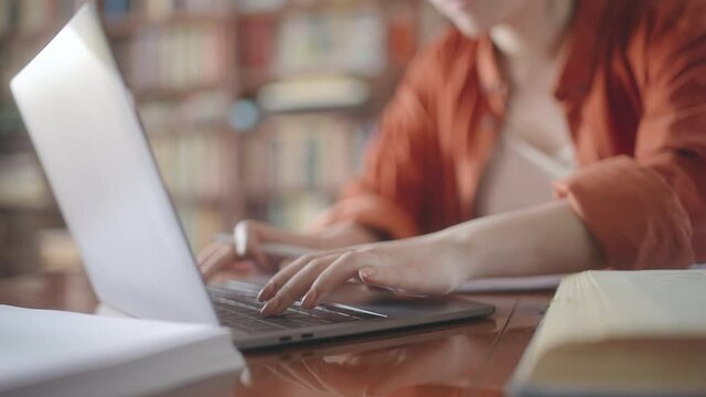 Female student doing research in a library, typing on laptop, reading books