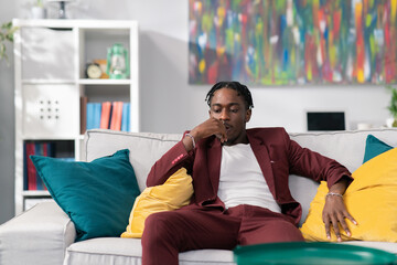 A thoughtful handsome man sits on couch, ponders a new project, dark-skinned businessman dressed in a maroon suit relaxes on sofa in room, thinks about work, money, business of the company