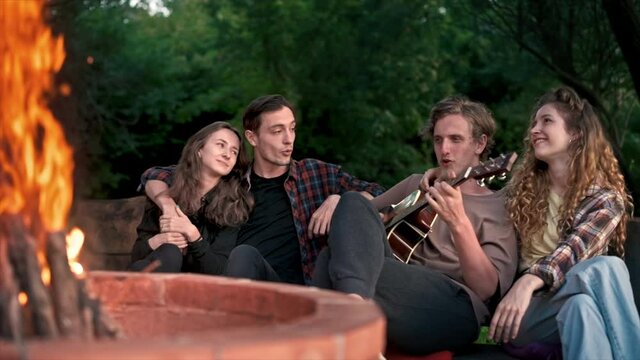A group of happy young friends near a campfire at glamping, night. Two men and women. Playing guitar. Slow motion