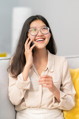 Portrait of laughing woman of Asian beauty, wearing beige clothes, shirt, dark hair eyes, silver round glasses, snow-white smile, eyes looking up, wide smile, talking on phone, calling family, friends