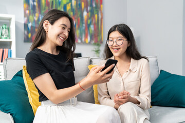Two adult pretty friends of Asian Korean beauty spend an afternoon on the living room couch, talking, the woman shows vacation photos on her phone, they look together, smile, comment