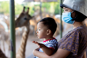 Happy mother wearing mask and Little kid boy watching giraffe in zoo. Happy child having excited...