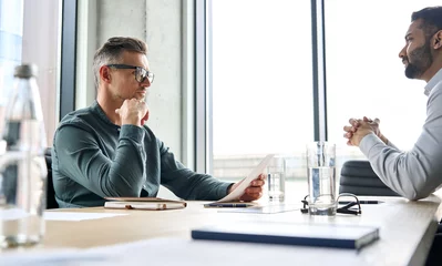Foto op Plexiglas Serious focused mature businessman employer hr or client holding cv or contract listening manager, applicant during job interview, two diverse ethnic partners having negotiations at office meeting. © insta_photos