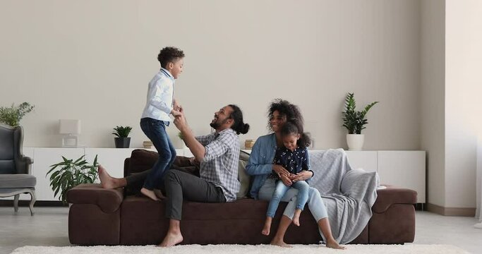 Full length joyful young african ethnicity couple parents having fun with cute little kids siblings in modern renovated apartment. Happy mixed race biracial family playing together in living room.
