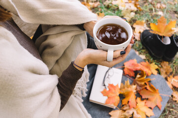 Photo of female in brown sweater and beige scarf warming hands on a cup with tea. Orange maple leaf and book lying on ground. Hot beverage for autumn days. Top view. Soft focus.