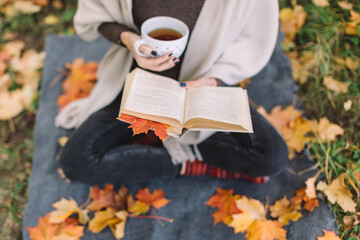 Woman in black jeans, brown sweater and beige scarf reading book and drink tea. Cup with warming beverage. Fallen maple leaves underfoot. Soft focus on leaf inside book. Autumn mood.